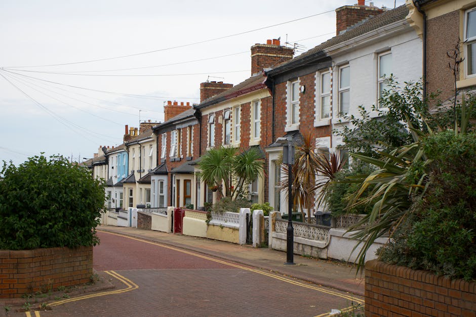 A row of terraced houses along a quiet residential street, featuring brick facades with varied colors and architectural details. In the foreground, a small garden bed contains lush green shrubs and a 